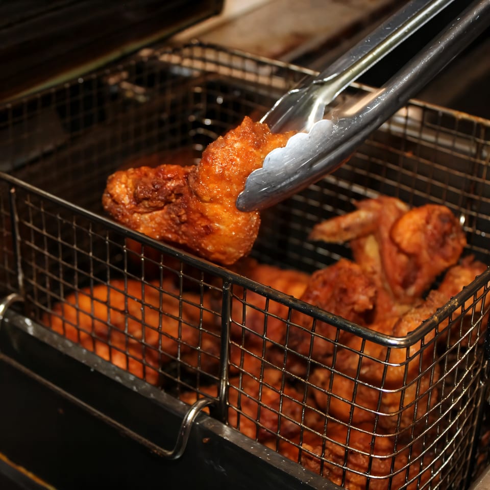 Tongs lift a piece of golden-brown fried chicken from a deep fryer basket at Ellenville Spice Fusion.