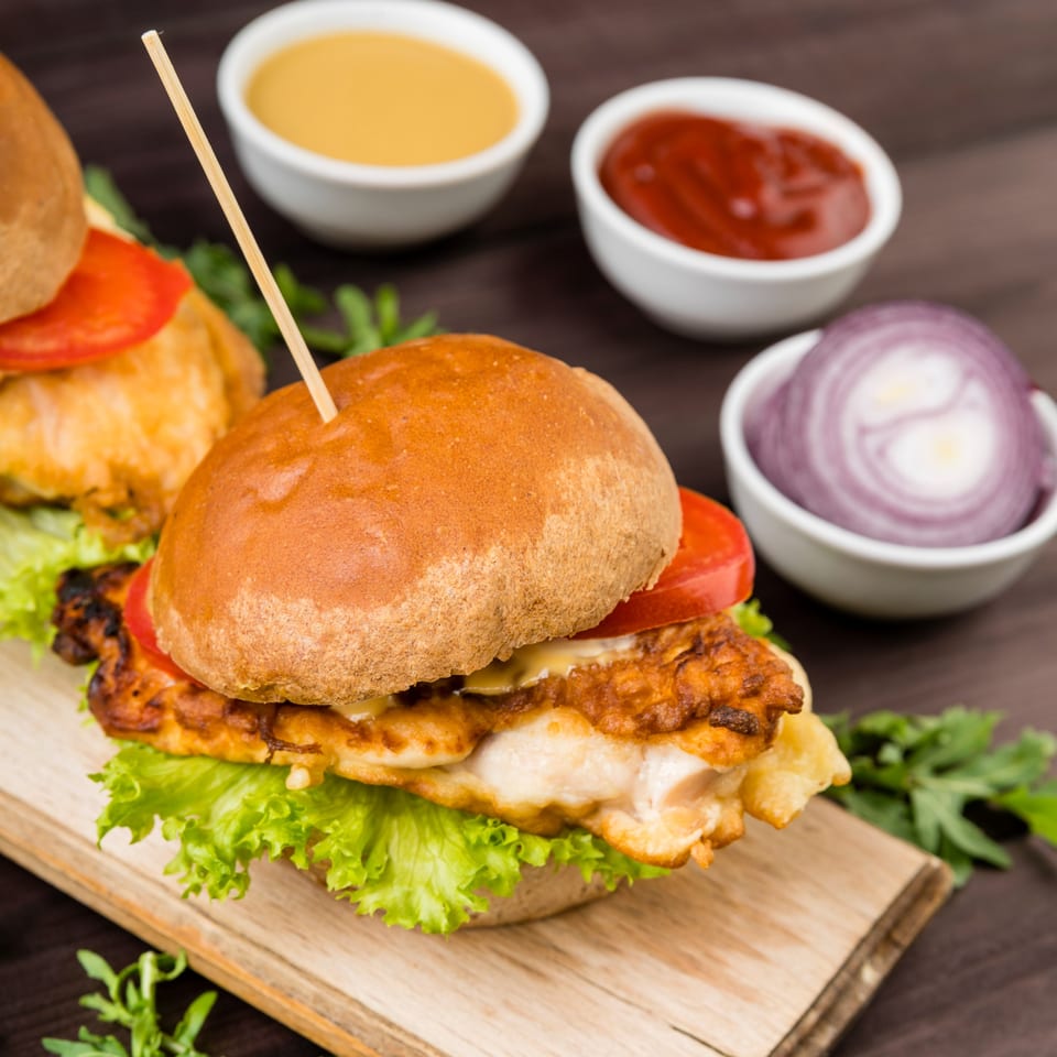 Two Ellenville Spice Fusion fried chicken sliders with lettuce, tomato, and sauces on a wooden board.