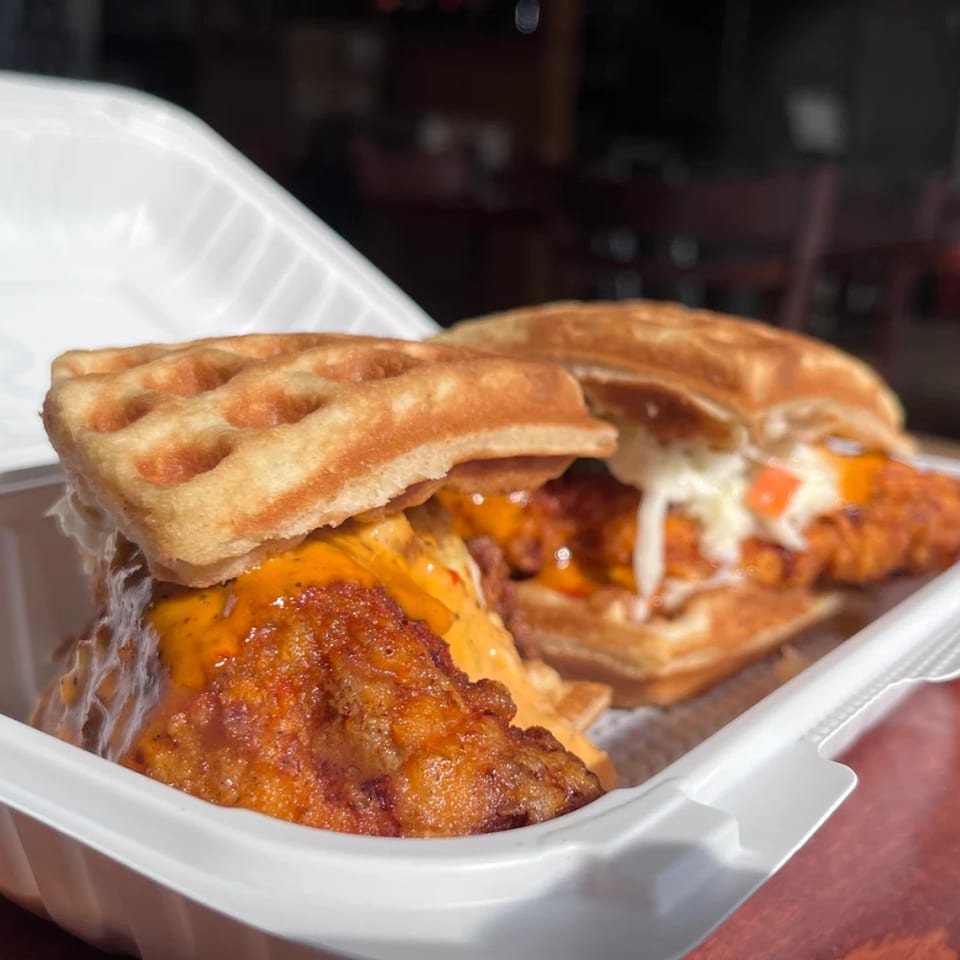 Two Ellenville Spice Fusion fried chicken and waffle sandwiches in a white takeout container.